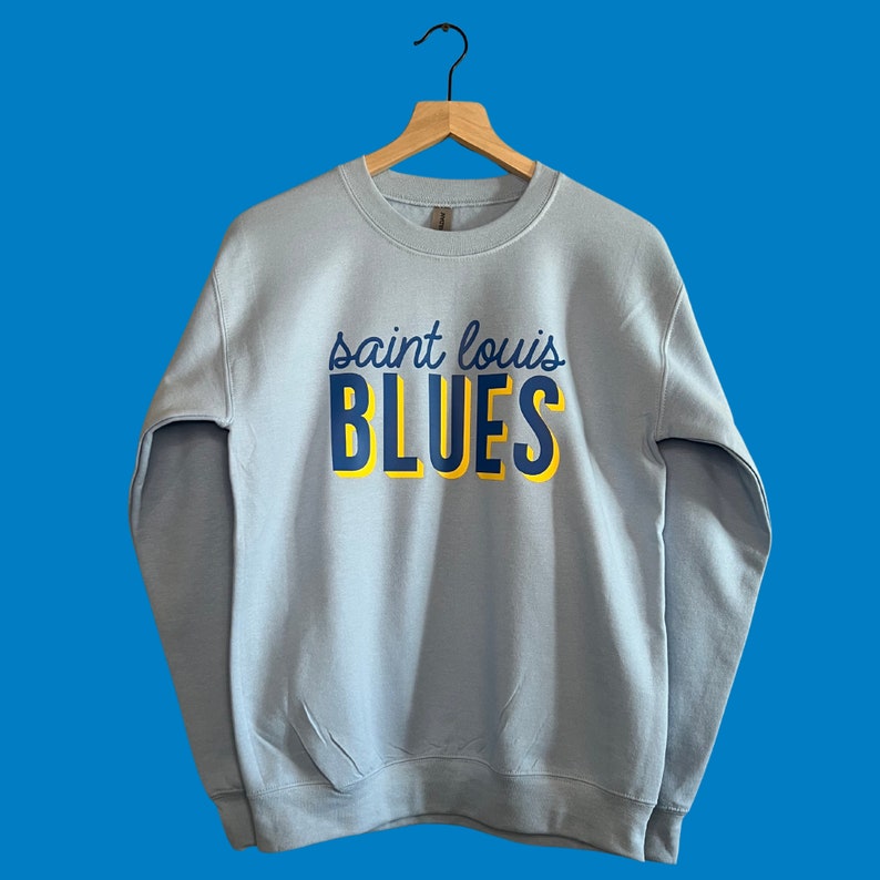 May include: A light blue sweatshirt with the words "saint louis BLUES" printed across the front in navy blue and yellow. The sweatshirt is hanging on a wooden hanger against a blue background.
