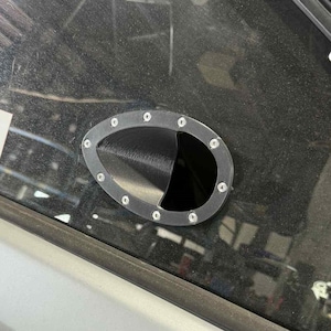 以下が含まれることがあります： 暗い内装の黒い楕円形の車の通気口。通気口はシルバーのトリムで囲まれ、小さなシルバーのボルトで固定されています。通気口は、暗い、おそらく黒い表面に取り付けられています。