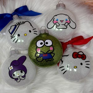 May include: Five glittery Christmas ornaments featuring cartoon characters. The ornaments include Hello Kitty, My Melody, Keroppi, and Cinnamoroll. Each ornament has a silver cap and is adorned with a ribbon in various colors.