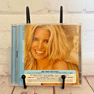 May include: A blue and white CD case with a photo of Jessica Simpson on the front. The case is for her album "In This Skin" and includes a Wal-Mart exclusive bonus DVD. The text on the case reads "New Expanded Version of Jessica's Hit Album Contains the Smash Hit "With You" + Three New Songs - "Take My Breath Away" "Angels"  Bonus DVD with Jessica's Wedding Footage and Scenes from Her Hit TV Show "The Newlyweds" Win a Trip to Meet Jessica Simpson!"