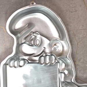 May include: A silver-colored Smurf-shaped cake pan. The pan features a smiling Smurf holding a rectangular sign. The pan has a loop at the top for hanging. The pan is made of metal and has a shiny finish.