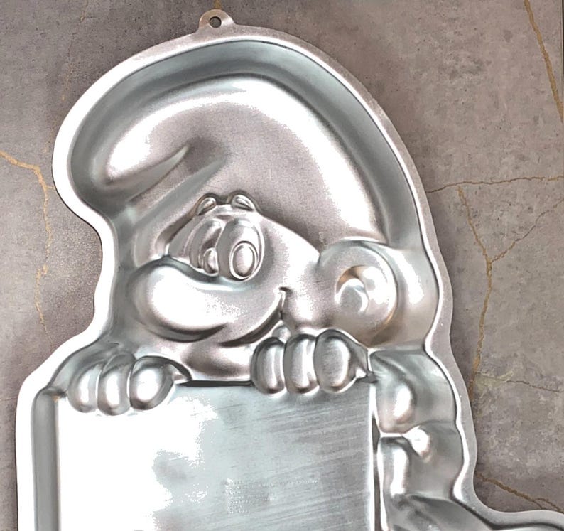 May include: A silver-colored Smurf-shaped cake pan. The pan features a smiling Smurf holding a rectangular sign. The pan has a loop at the top for hanging. The pan is made of metal and has a shiny finish.
