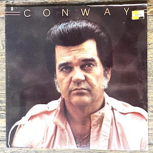 May include: A vinyl record album cover featuring a portrait of Conway Twitty. The text CONWAY is printed in white at the top. The subject has dark hair and sideburns, wearing a light pink collared shirt. A small yellow Venture sticker is visible.