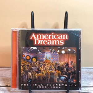May include: A CD case for the "American Dreams" original soundtrack, 1963-1964. The cover features the title in red and white, with a black and white image of a crowd watching a performance. The CD is in a clear plastic case.