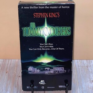 May include: A VHS tape for Stephen King's "The Tommyknockers" with a dark background and a green and white graphic. The text reads "A new thriller from the master of horror." and "You Can't Run. You Can't Hide. You Can Only Become...One Of Them."