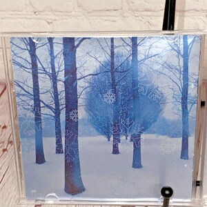May include: A CD case with a wintery scene of trees and snowflakes. The background is a light blue color and the trees are a darker blue. The snowflakes are white and are scattered throughout the image.