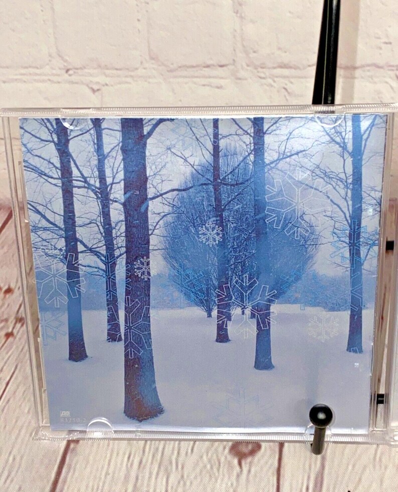 May include: A CD case with a wintery scene of trees and snowflakes. The background is a light blue color and the trees are a darker blue. The snowflakes are white and are scattered throughout the image.