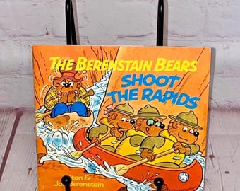 May include: A vintage children's book titled "The Berenstain Bears Shoot the Rapids" featuring illustrations of the bear family in a canoe going over rapids. The book cover is orange, blue, and green with a white star on the canoe.