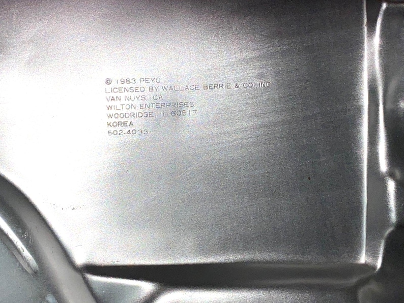 May include: Close-up of a silver metal baking pan with text that reads "&copy; 1983 PEYO LICENSED BY WALLACE BERRIE & CO., INC VAN NUYS, CA WILTON ENTERPRISES WOODRIDGE, IL 60517 KOREA 502-4033."