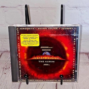 Puede incluir: Una caja de CD para la banda sonora de la película Armageddon. La caja es negra con una imagen de fondo roja y naranja de un planeta. El título "Armageddon: The Album" está impreso en blanco en la caja. El álbum presenta artistas como Aerosmith, Jon Bon Jovi y Journey.