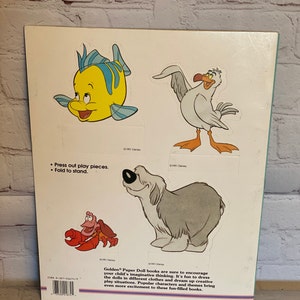 May include: A book with a white background and colourful illustrations of characters from the Disney film The Little Mermaid. The book includes instructions to press out the play pieces and fold to stand. The text on the back of the book reads "Golden Paper Doll books are sure to encourage your child's imaginative thinking. It's fun to dress the dolls in different clothes and dream up creative play situations. Popular characters and themes bring even more excitement to these fun-filled books."