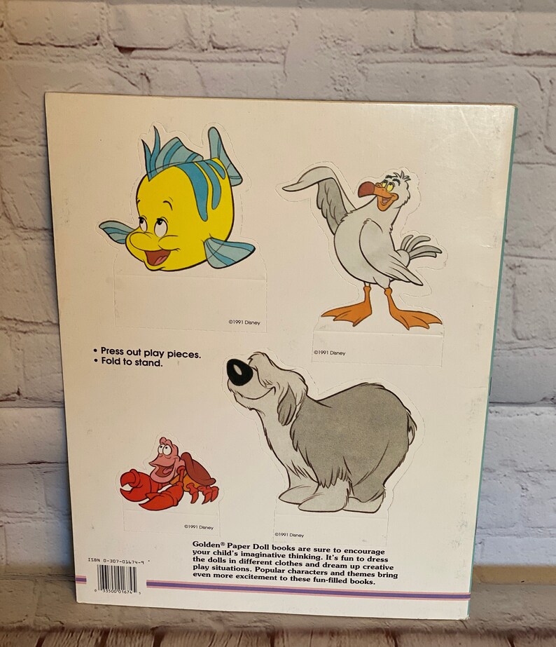 May include: A book with a white background and colourful illustrations of characters from the Disney film The Little Mermaid. The book includes instructions to press out the play pieces and fold to stand. The text on the back of the book reads "Golden Paper Doll books are sure to encourage your child's imaginative thinking. It's fun to dress the dolls in different clothes and dream up creative play situations. Popular characters and themes bring even more excitement to these fun-filled books."