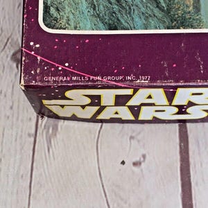 以下が含まれることがあります： ヴィンテージのスターウォーズの箱の一部で、黄色のブロック文字で「STAR WARS」と書かれています。箱は紫色の背景に白い斑点があり、「GENERAL MILLS FUN GROUP, INC. 1977」と書かれています。