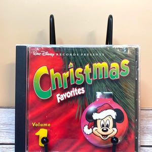 May include: A Walt Disney Records Presents Christmas Favorites Volume 1 CD case. The cover features a red background, green pine needles, and a pink Christmas ornament with a graphic of Mickey Mouse wearing a Santa hat.
