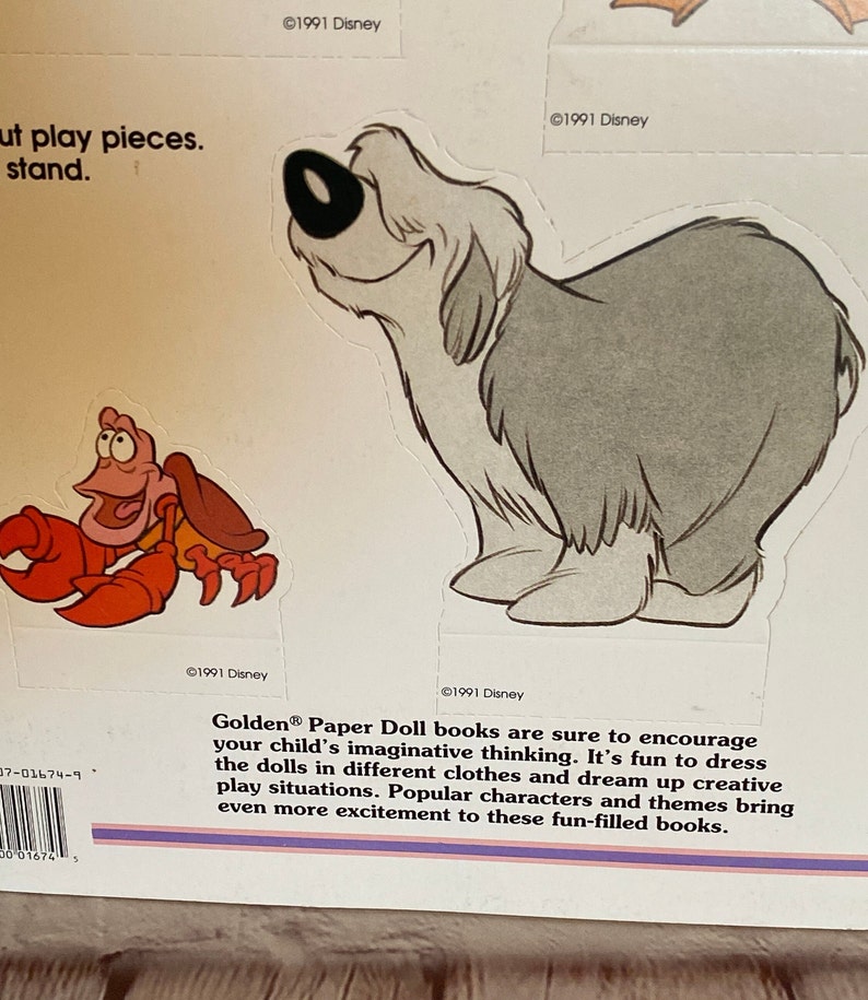 May include: A vintage Golden Paper Doll book featuring a red crab and a grey dog. The book cover includes the text "Golden Paper Doll books are sure to encourage your child's imaginative thinking. It's fun to dress the dolls in different clothes and dream up creative play situations. Popular characters and themes bring even more excitement to these fun-filled books."