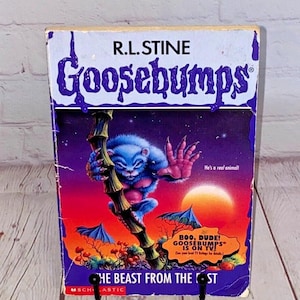Puede incluir: Un libro de bolsillo vintage titulado "Goosebumps: The Beast From The East" de R.L. Stine. La portada presenta una criatura azul y peluda con un pecho rojo, agarrada a un tallo de bambú contra un fondo de puesta de sol. El libro tiene un borde púrpura y el texto "BOO, DUDE! GOOSEBUMPS IS ON TV!"