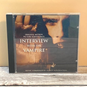 May include: A CD case featuring the "Interview with the Vampire" original motion picture soundtrack. The cover art displays a close-up of a man's face, with the title text in white and the composer's name at the bottom.