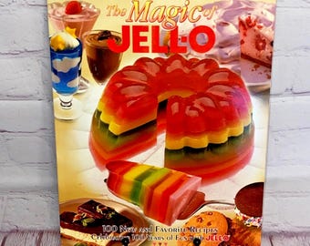 Op de afbeelding: Een kookboek getiteld "The Magic of JELLO" met een kleurrijk gelaagd JELLO-dessert op de omslag. Het boek bevat 100 nieuwe en favoriete recepten en viert 100 jaar plezier met JELLO.