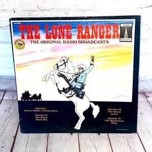 Puede incluir: Una portada de álbum de radiodifusión vintage de "The Lone Ranger". La portada presenta a un vaquero sobre un caballo blanco encabritado, con el título en rojo y las palabras "The Original Radio Broadcasts". El álbum es un lanzamiento de Wonderland Records.