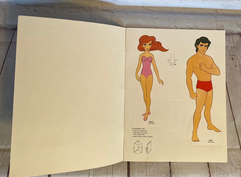 May include: A paper doll set featuring Ariel and Eric from the Disney movie The Little Mermaid. The dolls are printed on cardstock and are ready to be cut out and assembled. Instructions for assembling the dolls are included on the page.
