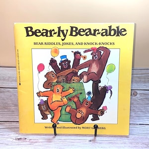 Puede incluir: Un libro infantil amarillo titulado Bear-ly Bear-able: Bear Riddles, Jokes, and Knock-Knocks de Mort Gerberg. La portada presenta una colorida ilustración de varios osos pardos jugando con globos, sombreros de fiesta e instrumentos alrededor de un barril.