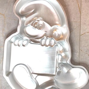 May include: A silver-colored Smurf-shaped cake pan. The pan features the character holding a blank sign. The pan has a loop at the top for hanging. The pan is made of metal and is designed for baking cakes.