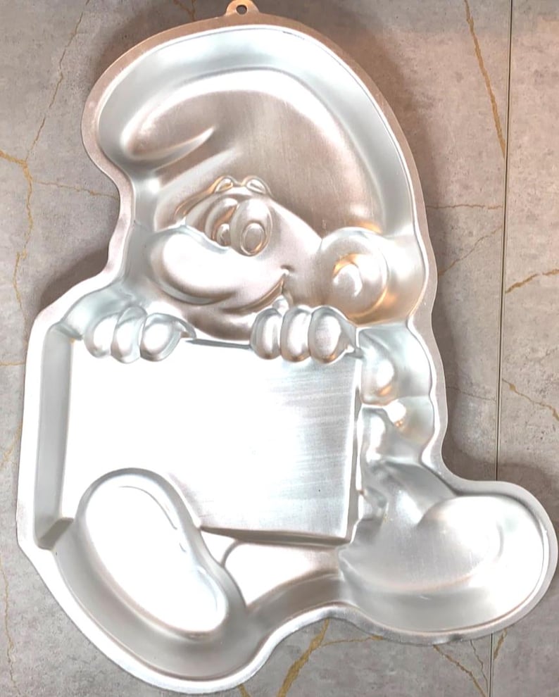 May include: A silver-colored Smurf-shaped cake pan. The pan features the character holding a blank sign. The pan has a loop at the top for hanging. The pan is made of metal and is designed for baking cakes.