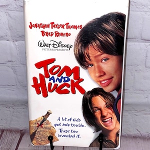 May include: A white VHS tape for the Disney movie "Tom and Huck" featuring Jonathan Taylor Thomas and Brad Renfro. The title is in red, with images of the actors. The tape is held by a black metal stand.