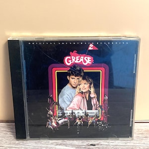 May include: A CD case featuring the "Grease 2" soundtrack. The cover art showcases a couple in a romantic embrace, set against a backdrop of a retro-style frame. The title "Grease" is prominently displayed, with the number "2" and the words "Original Soundtrack Recording" visible.