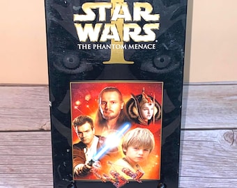 Könnte beinhalten: Eine schwarze VHS-Kassette von Star Wars: Die dunkle Bedrohung. Das Cover zeigt das Star Wars-Logo und Bilder von Charakteren aus dem Film. Die Kassette ist mit "Digitally Superior Bound" und "THX Mastered for Picture Quality" beschriftet.