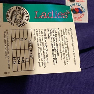 May include: A purple Fruit of the Loom t-shirt with a white tag that says "Ladies'" and has a size chart. The tag also says "Unconditionally Guaranteed" and includes information about the brand and its products.