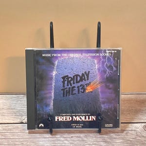 May include: A Friday the 13th The Series music CD in a clear plastic jewel case. The cover art features a stone tombstone against a dark purple stormy sky with lightning. Text reads Music From The Original Television Scores, Fred Mollin, and Over 70 min. of music.
