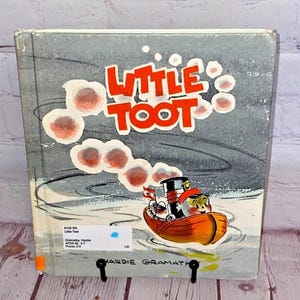 May include: A vintage children's book cover featuring a red and black tugboat named "Little Toot" with a smiling face. The boat is sailing on a blue body of water with white waves and is leaving a trail of red smoke.