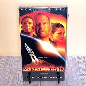 Puede incluir: Una cinta VHS de la película "Armageddon" con Bruce Willis. La portada muestra una escena ardiente con un cohete y el título de la película. La parte inferior de la caja dice "For Love. For Honor. For Man."