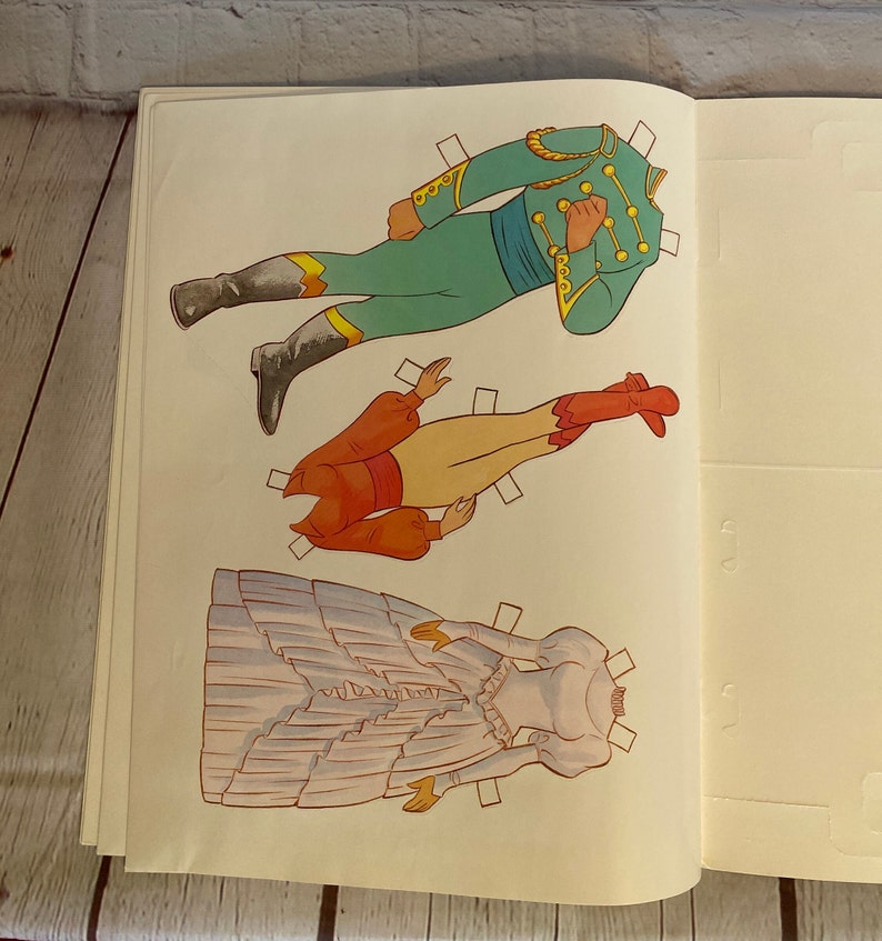 May include: A vintage paper doll set featuring a couple in traditional attire. The woman wears a white gown with a ruffled skirt and a red bodice. The man wears a green jacket with gold trim and red trousers. The paper dolls are ready to be cut out and played with.