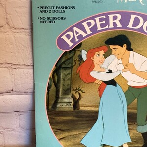 May include: A blue and pink paper doll book featuring Ariel and Prince Eric from Disney's The Little Mermaid. The book includes precut fashions and two dolls. The text on the cover reads "The Little Mermaid Paper Dolls".