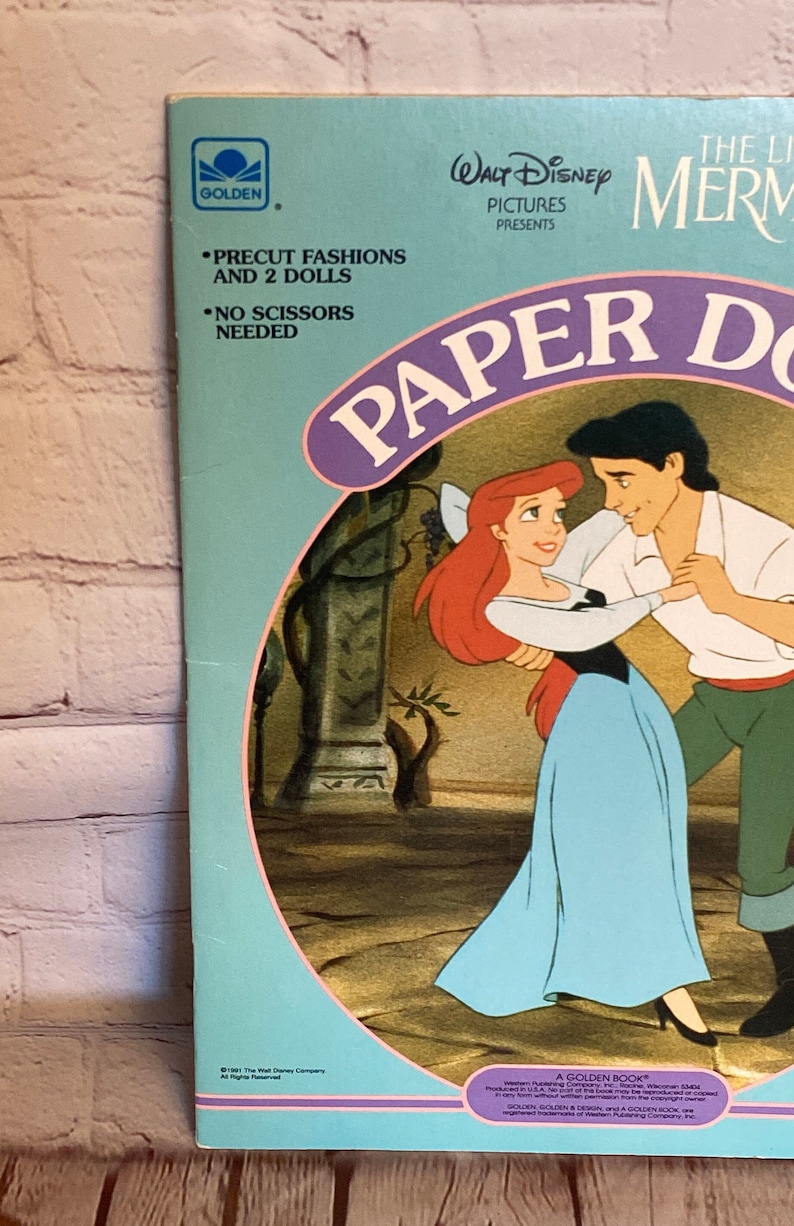 May include: A blue and pink paper doll book featuring Ariel and Prince Eric from Disney's The Little Mermaid. The book includes precut fashions and two dolls. The text on the cover reads "The Little Mermaid Paper Dolls".