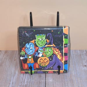 May include: A Halloween-themed napkin holder featuring a colorful illustration of a vampire, Frankenstein's monster, and a witch. The holder has a black background with stars and bats, and a black metal stand.