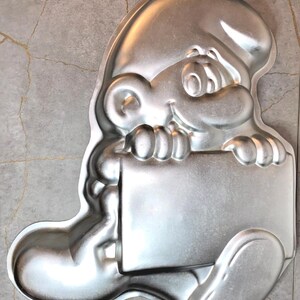 May include: A silver-colored metal cake pan in the shape of a cartoon character holding a blank sign. The pan has a loop for hanging and is designed for baking a shaped cake. The character has a smiling face and a large hat.