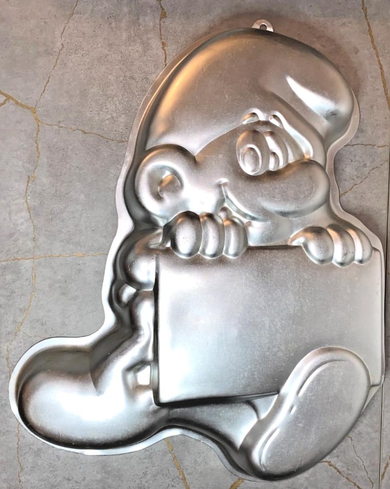 May include: A silver-colored metal cake pan in the shape of a cartoon character holding a blank sign. The pan has a loop for hanging and is designed for baking a shaped cake. The character has a smiling face and a large hat.