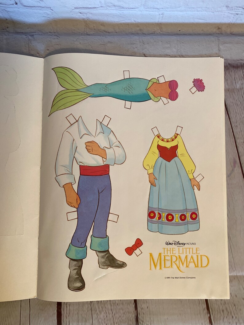 May include: A paper doll set featuring Ariel, Prince Eric, and a mermaid tail from the Disney film The Little Mermaid. The paper dolls are ready to be cut out and played with.