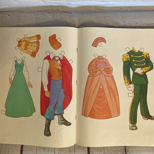 May include: A vintage paper doll set featuring a prince and princess. The prince is wearing a red cape and blue trousers, while the princess is wearing a pink gown. The set includes various clothing options for both characters.