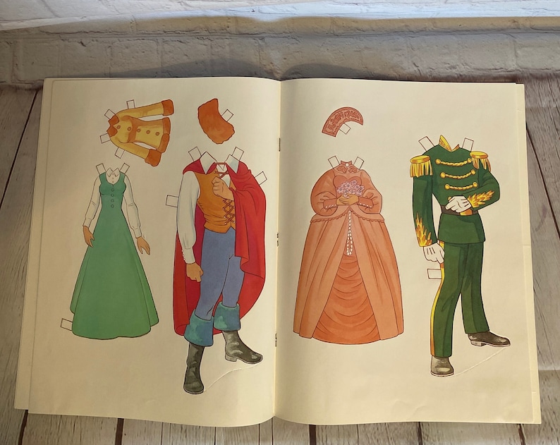 May include: A vintage paper doll set featuring a prince and princess. The prince is wearing a red cape and blue trousers, while the princess is wearing a pink gown. The set includes various clothing options for both characters.