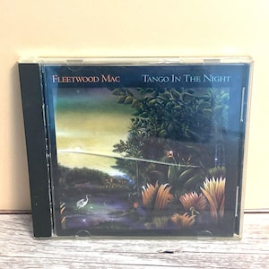 May include: A CD case featuring the album "Tango In The Night" by Fleetwood Mac. The cover art depicts a lush, tropical landscape with a body of water, plants, and a bird. The text "Fleetwood Mac" and "Tango In The Night" are visible.