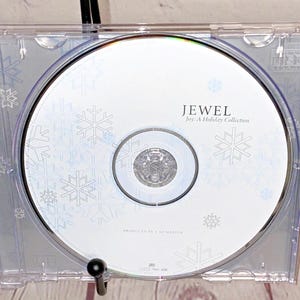 May include: A white CD with a snowflake design. The text "Jewel Joy: A Holiday Collection" is printed on the CD. The text "Produced by  &  The  Mardin" is printed on the CD.