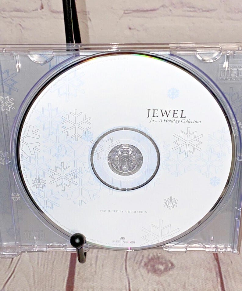 May include: A white CD with a snowflake design. The text "Jewel Joy: A Holiday Collection" is printed on the CD. The text "Produced by  &  The  Mardin" is printed on the CD.