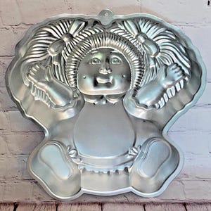 Puede incluir: Un molde para pastel de metal plateado con forma de muñeca con coletas. La muñeca lleva un vestido y tiene una cabeza grande con una cara sonriente.