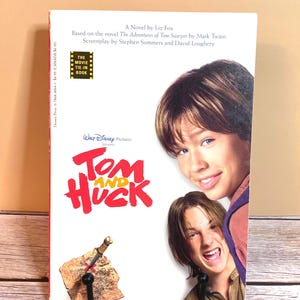 May include: A paperback book titled "Tom and Huck" with a movie tie-in label. The cover features two young men and the title in red and white. The book is propped up on a black hook.