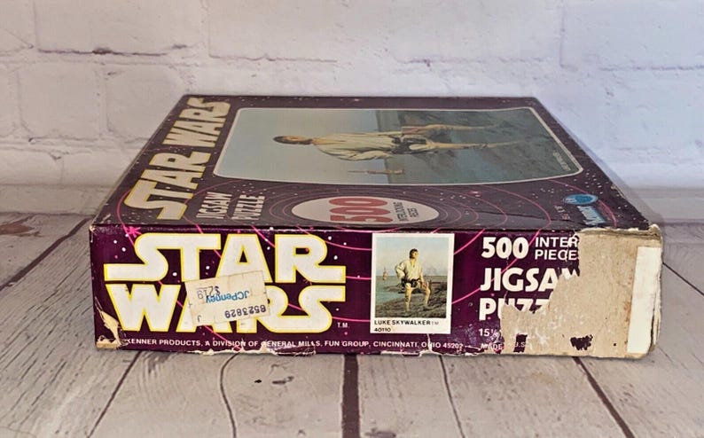 以下が含まれることがあります： ヴィンテージのスターウォーズ500ピースジグソーパズルボックス。ボックスは主に紫で、スターウォーズのロゴは黄色と白です。ボックスには、ルーク・スカイウォーカーの画像と「500 Piece Jigsaw Puzzle」のテキストが表示されています。