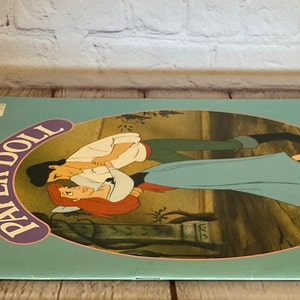 May include: A vintage Disney's The Little Mermaid paper doll book with a colourful cover featuring Ariel and Prince Eric. The cover has the text "Paper Doll" in a purple and white circle and "Disney's The Little Mermaid" in a blue and white rectangle.